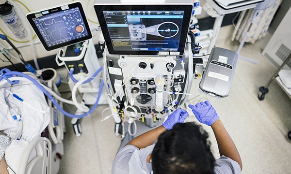 A nurse consults two screens of digital information about a patient lying on a hospital bed