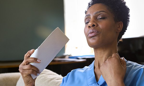 A nurse in scrubs sits while taking a break, looking tired and fanning her face with her eyes closed as she struggles with symptoms of menopause during a shift