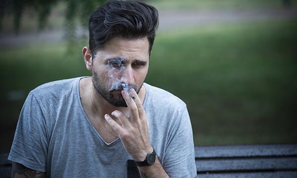 A sad-looking young man sits on a park bench alone smoking a cigarette