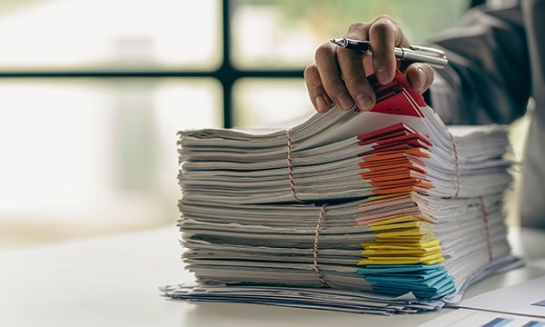 Alt text pic 1: A person’s hand sits atop a stack of papers colour-coded as if for case files, suggesting the backlog of cases at the nursing regulator, the NMC