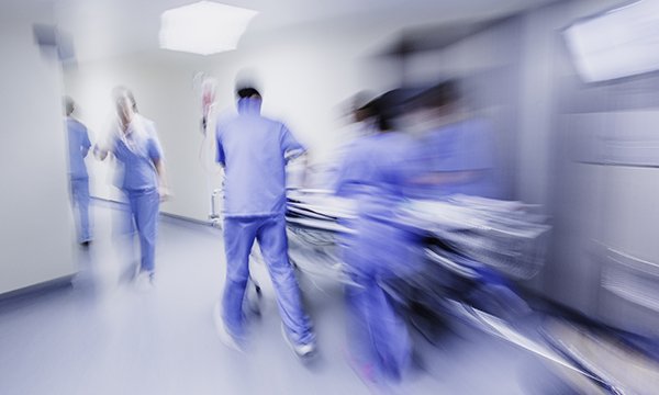 Blurred image of healthcare professionals moving at high speed with a hospital trolley along a corridor, suggesting the time and workload pressures facing emergency department staff 