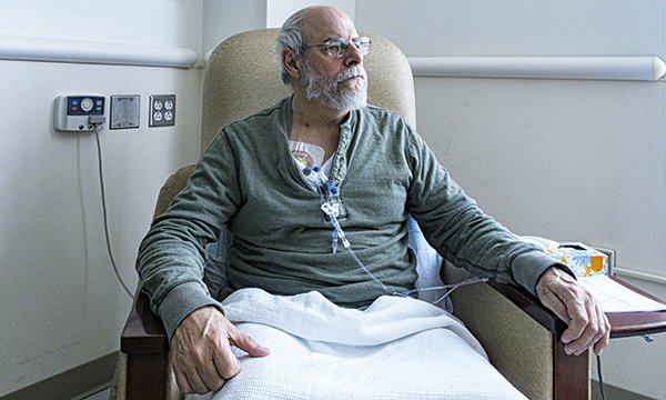An older person sitting on a comfortable chair who is hooked up to a several tubes on his chest, with a light blanket on his lap