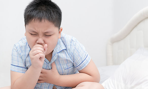 A young boy coughs into his right hand while holding his chest with the other