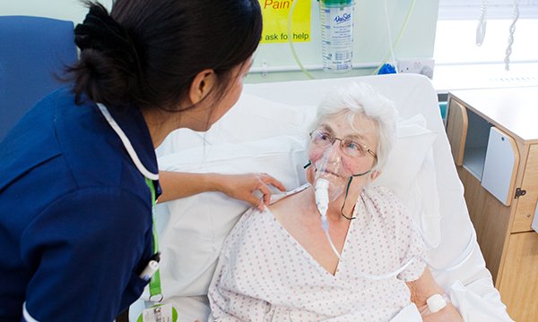 nurse who appears to be of Filipino origin leans towards female inpatient who is having oxygen therapy
