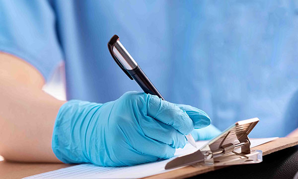 A nurse in uniform holding a pen and writing in a patient’s notes while wearing disposable gloves, highlighted in the Be PPE Free campaign as an inappropriate use of PPE