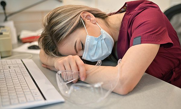 A nurse wearing a mask and resting her head on a desk, alongside a computer.