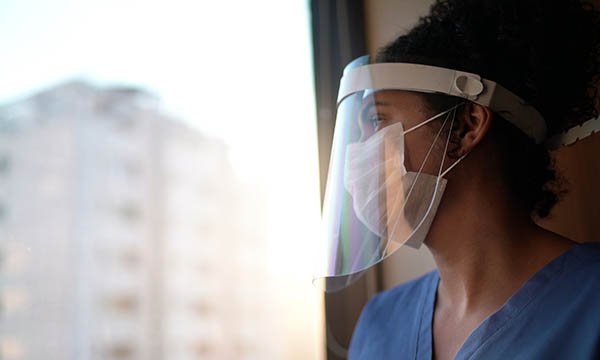 A nurse in PPE visor and facemask staring out a hospital window