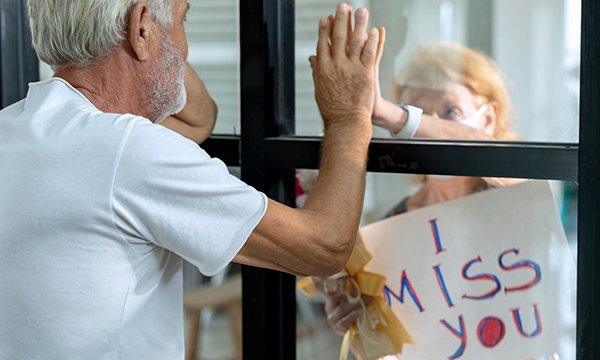 Picture shows person looking through a care home window at a family member with a placard saying: 'I miss you'