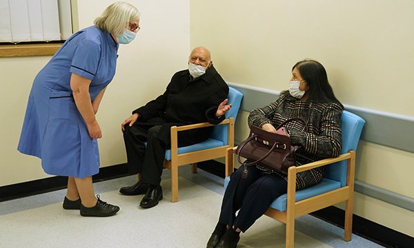 Nurse wearing a facce mask talking to two patients in a clinic waiting room, both wearing masks