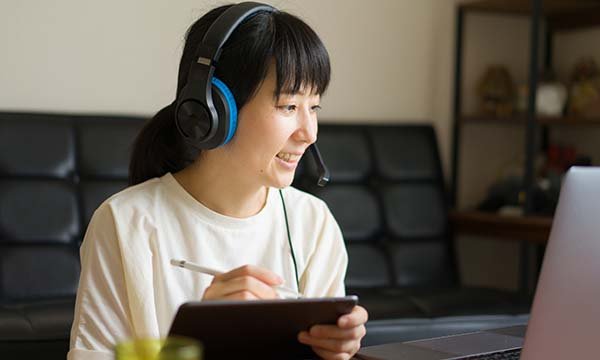Picture sows a smiling young woman wearing headphones and taking notes as she looks at a computer