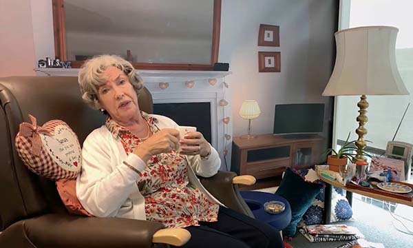 A still from The Street where an actor plays an older woman sitting in her living room drinking tea