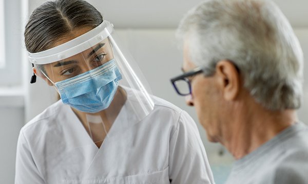 A nurse in a face mask and visor talks to an older patient