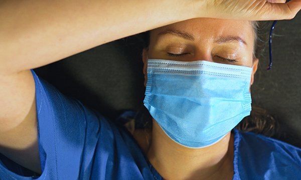 A nurse in PPE looking exhausted and overheated, with her arm resting on her forehead