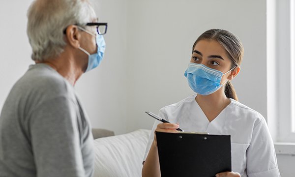 Masked older man having a consultation with a masked nurse with a clipboard and pen
