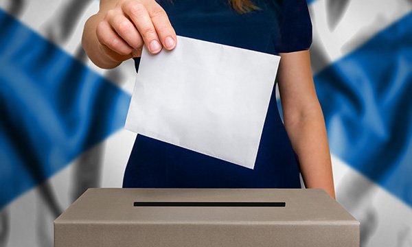 Photo of a voting slip being placed into a ballot box with a Scottish flag in the background