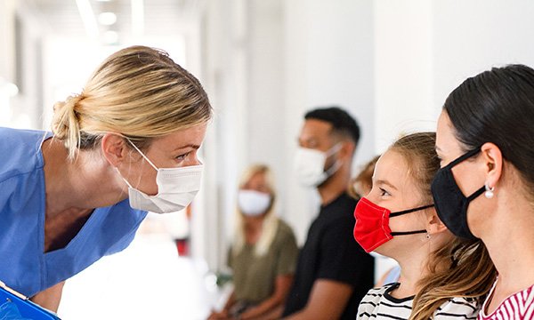 Standing nurse leans down to talk to a girl and woman seated, all wearing masks