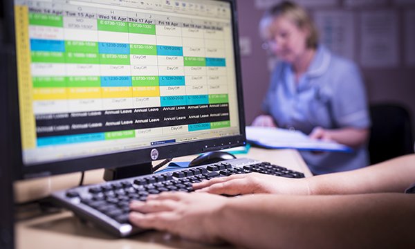 Picture shows a computer screen displaying a work rota, with a nurse in the background