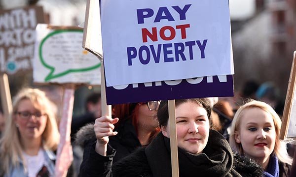 A protest march with a woman holding a placard saying 'Pay not poverty'