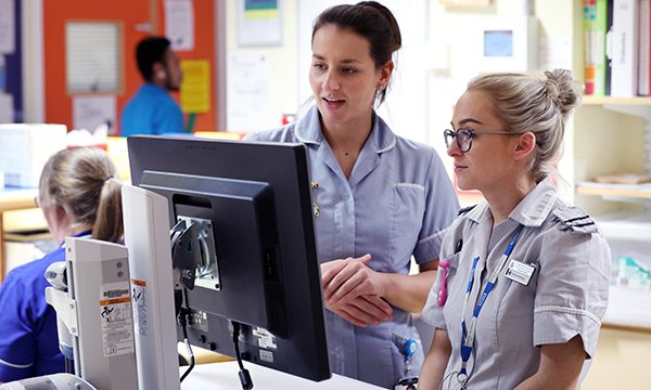 A nurse and nursing student on clinical placement talk at ward computer as nurses debate idea of reducing practice hours