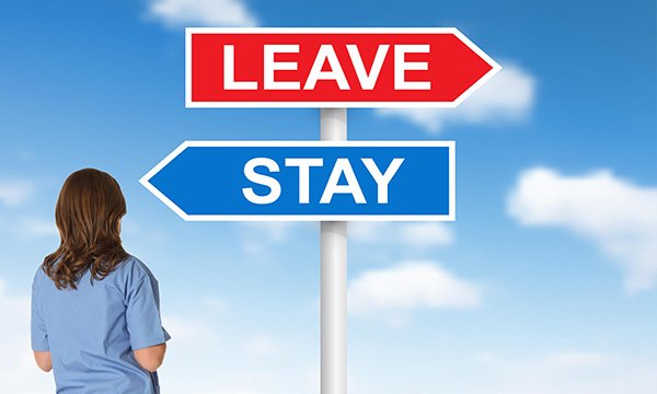 Nurse standing in front of a street sign with red ‘leave’ sign pointing one way and blue ‘stay’ sign the other way, against a background of a blue sky with clouds, representing the choice nurses face over remaining in or leaving the profession