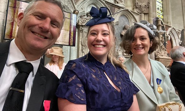 Nurse Emily Regan smiles, seated in Westminster Abbey between Adam Hills and Claire Spencer