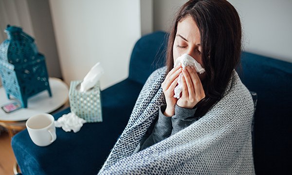 Woman on sofa, wrapped in blanket, blowing her nose and looking too ill to work