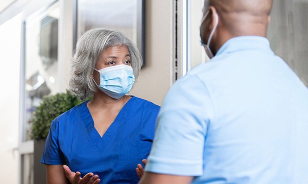 An older female nurse in uniform wearing a mask, talking to a male staff member