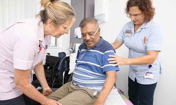 Two older nurses standing on either side of an older male patient, helping him, as he sits on a bed in a consultation room