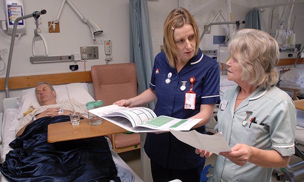 An older nurse talks to a younger nursing colleague on a ward, while a patient lies on a hospital bed in the background. Older nurses often bring wider knowledge and experience to their roles