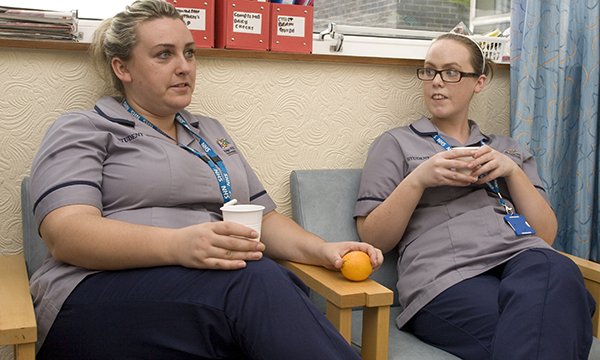 Two nurses taking a break in a work break room for staff, sitting in chairs and holding drinking cups