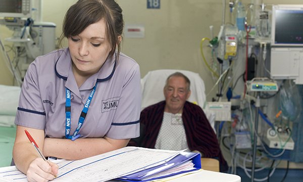 A nursing student filling in paperwork in the foreground as a patient sits in a chair next to his hospital bed