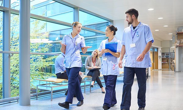 Three nurses in scrubs walk along a hospital corridor, talking animatedly