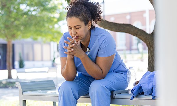 A nurse in blue scrubs sits on a bench outside her workplace, with her hands together and looking down as if she is feeling worried