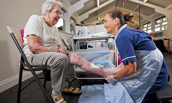 A patient and nurse smile at each other while the nurse kneels holding the patient’s leg, which has a newly applied dressing on the shin