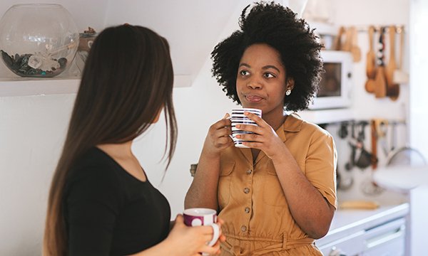 Two women talk to each other, both holding a cup of tea. Support from colleague can help individuals deal with imposter feelings