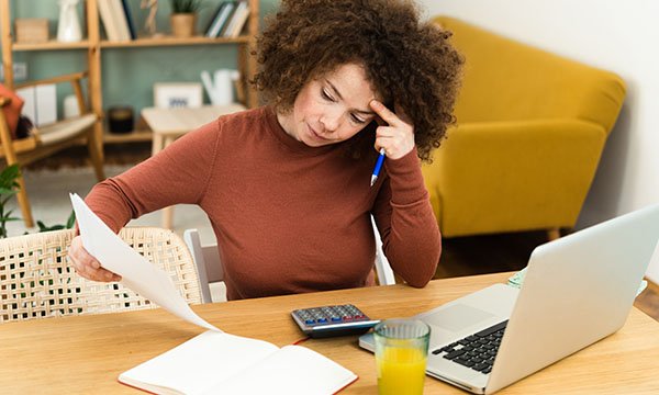 A woman sits at a table at home looking anxious with a calculator and laptop in front of her