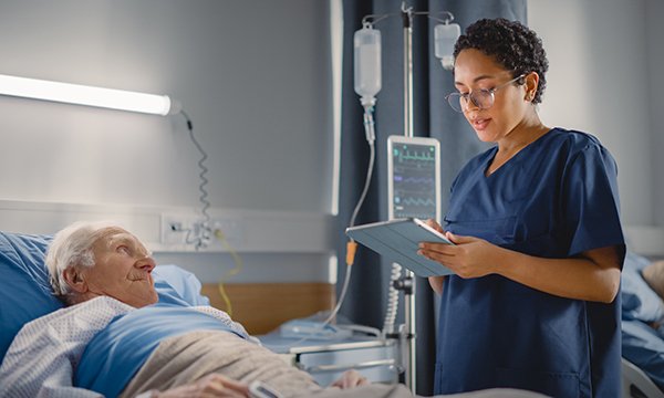 Nursing standing at the bedside of an older male patient making note on an electronic tablet