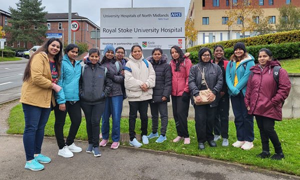 Twelve overseas nurse recruits stand and smile in front of the Royal Stoke University Hospital sign