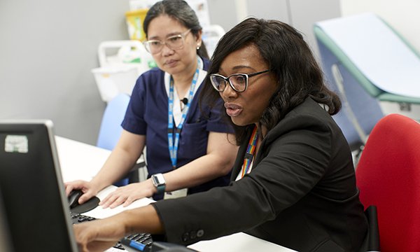 RCN Nurse of the Year 2023 Julie Roye, pictured siting at a desk at her workplace alongside colleague Jennifer Santos, pointing to a computer screen