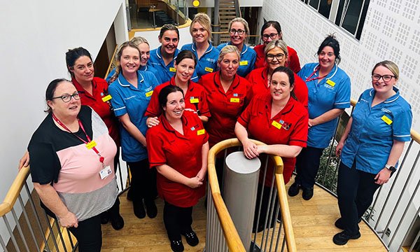 A photo of 15 members of the Arches district nursing team standing on a staircase and smiling up at the camera