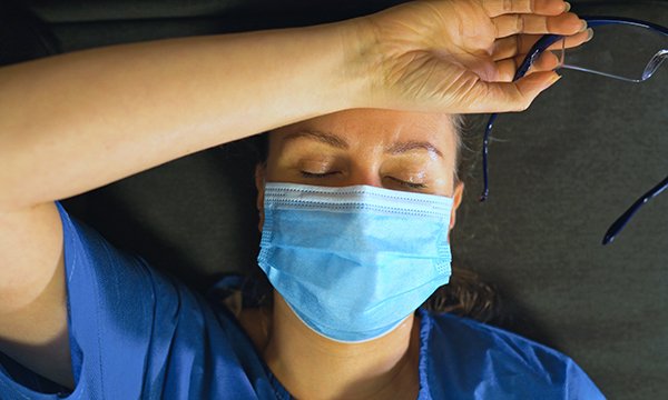 Photo of a nurse in a face mask lying on the floor with her eyes shut, glasses off and the back of her hand on her forehead