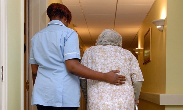 A nurse puts her hand on the back of an older woman to support her as she walks with the help of a walking frame