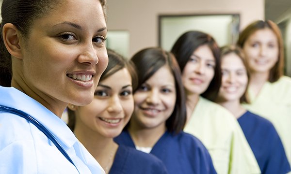 Six smiling nurses stand in a line, with the nearest one most in focus 