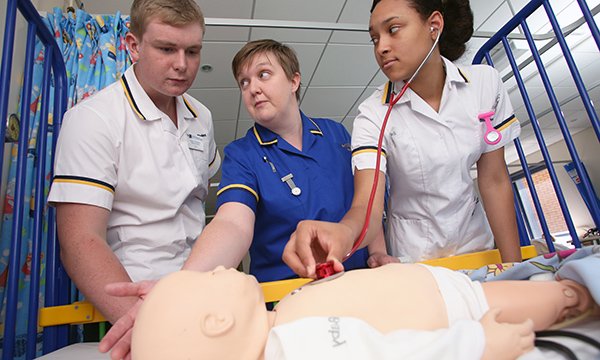 Nurse is flanked by two nursing students in simulated clinical learning setting. One student is listening to chest of manikin infant
