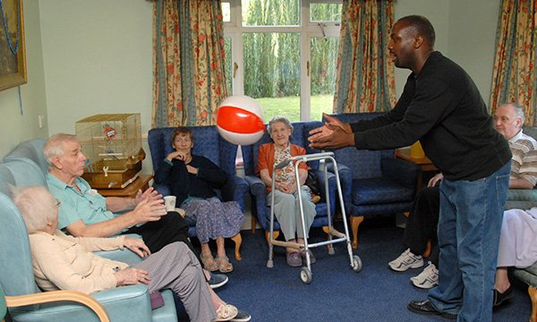 Picture shows a group of older people sitting in chairs while a younger man in the centre of the room throws one of them a beach ball. A four-year project will build data resource on treatment and services at care homes.