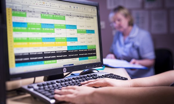 Picture shows the hands of a person looking at a computer screen that is displaying a work roster, with a medic in the background