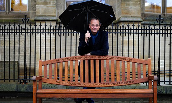 Picture shows broadcaster Mark Radcliffe standing next to a park bench in the grounds of the University of Manchester which bears his tribute to cancer research. 
