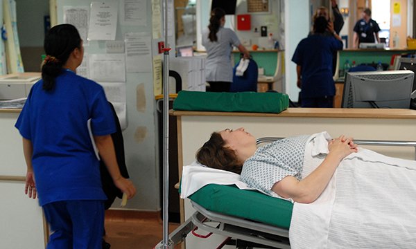 Picture shows a nurse standing next to a woman lying on a trolley in a hospital corridor. The official NHS count of 12-hour waits at emergency d­­epartments in England underestimates the actual figure, says the Royal College of Emergency Medicine.