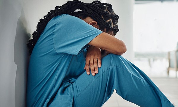 A female black nurse sitting with her back against a wall with knees raised, leaning forward and resting her head against her arms to indicate sadness or depression 