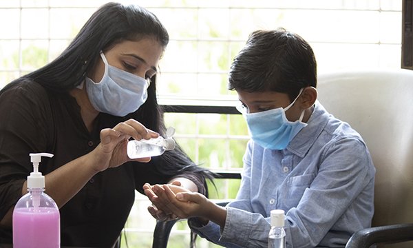 Picture of a woman showing a boy how to clean his hands with sanitiser. Both are wearing masks.